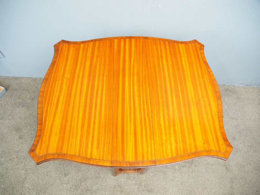 An Edwardian satinwood Sutherland table shown folded with curved drop-leaf sides, fretwork side supports, and brass castors. View III