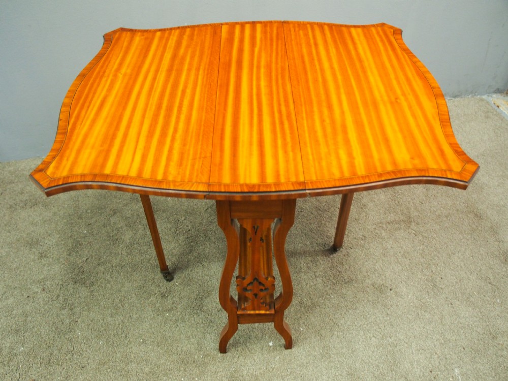 An Edwardian satinwood Sutherland table shown folded with curved drop-leaf sides, fretwork side supports, and brass castors. View IV