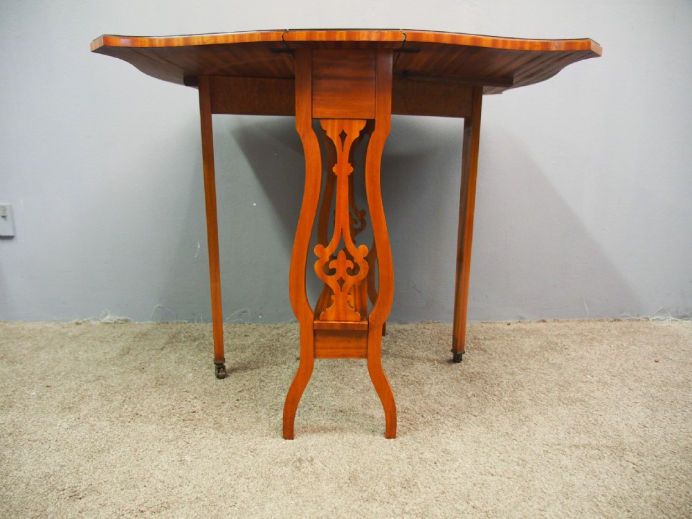 An Edwardian satinwood Sutherland table shown folded with curved drop-leaf sides, fretwork side supports, and brass castors. View VI