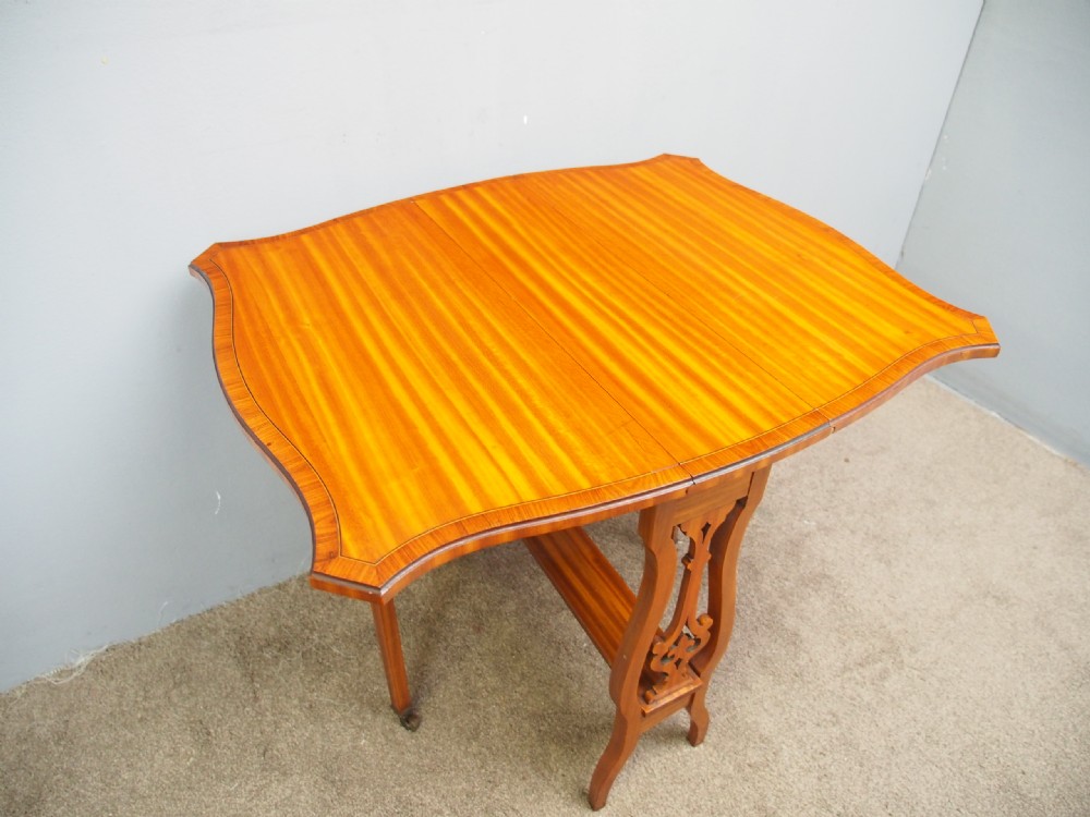 An Edwardian satinwood Sutherland table shown folded with curved drop-leaf sides, fretwork side supports, and brass castors. View VII