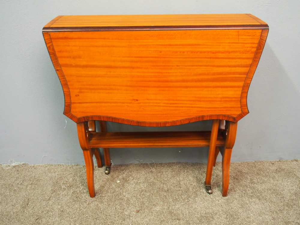 An Edwardian satinwood Sutherland table shown folded with curved drop-leaf sides, fretwork side supports, and brass castors. View VIII