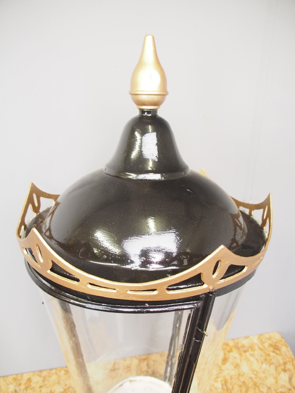 A tapered circular street lamp lantern featuring a black domed cap with a gold finial, gold filigree trim, and clear glass side panels. View IV