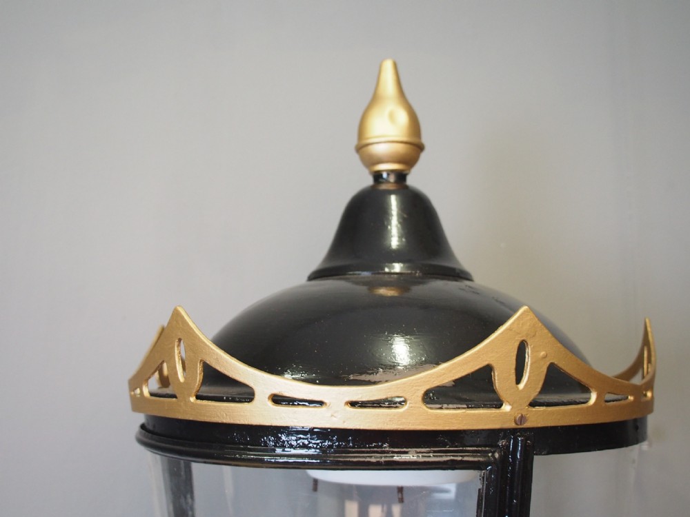 A tapered circular street lamp lantern featuring a black domed cap with a gold finial, gold filigree trim, and clear glass side panels. View V