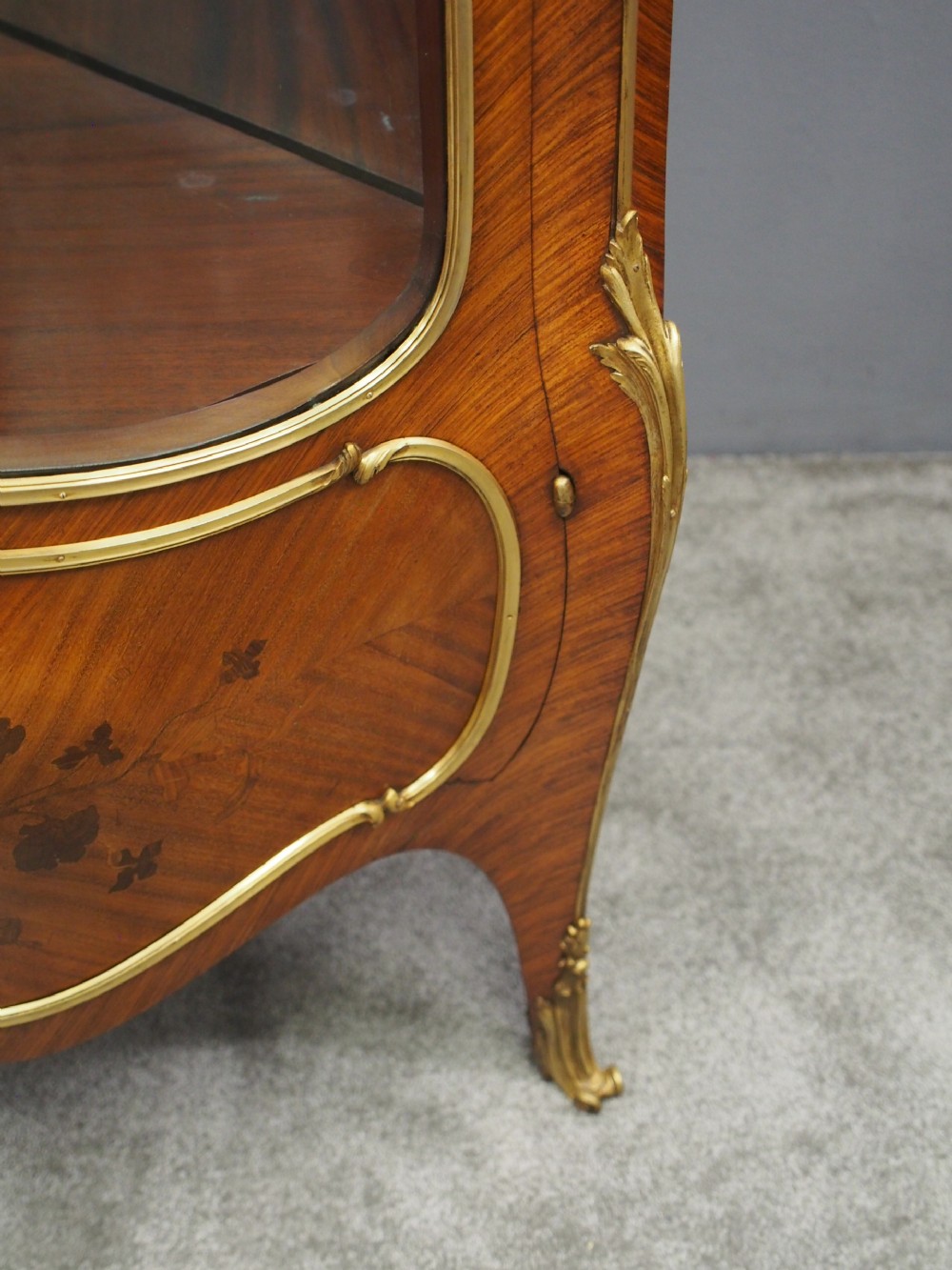 A Victorian corner vitrine in kingwood featuring curved glass doors, floral marquetry on the lower panel, and ornate brass ormolu mounts. View VI