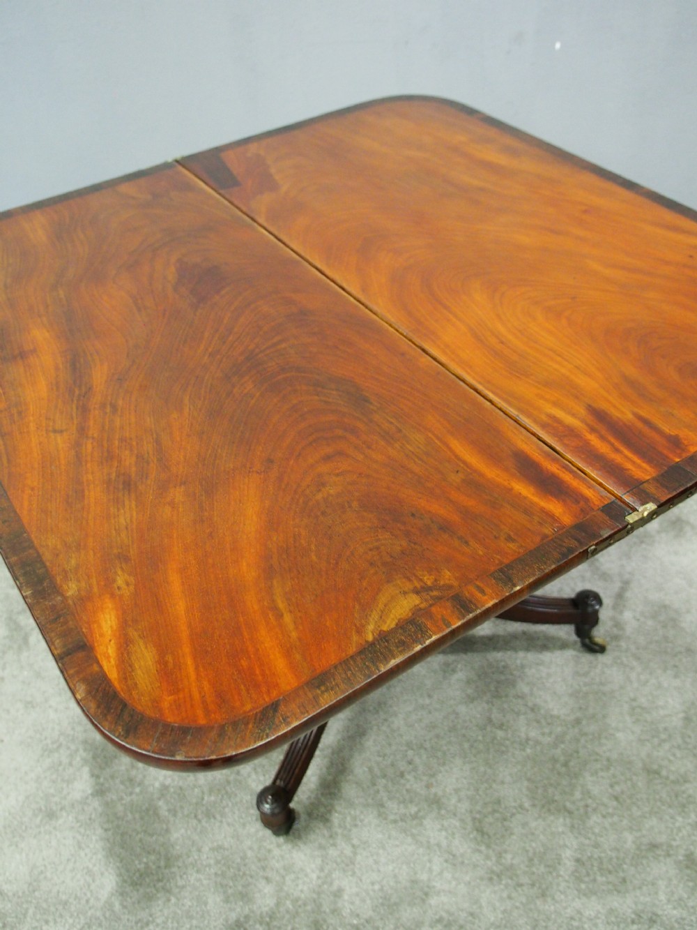 A Regency mahogany foldover card table featuring rosewood inlay along the apron, a turned central pedestal, and four swept legs with brass castors. View X