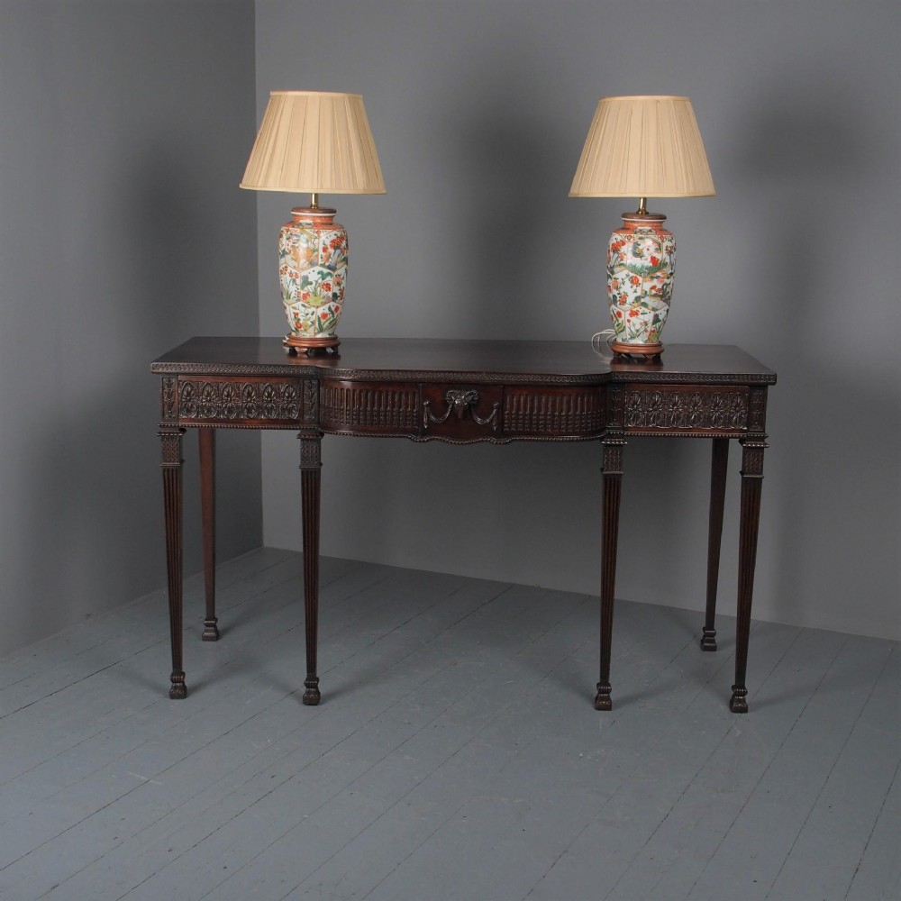 An Adam style carved dark mahogany serving table featuring fluted tapered legs, a central drawer, and detailed frieze carving.