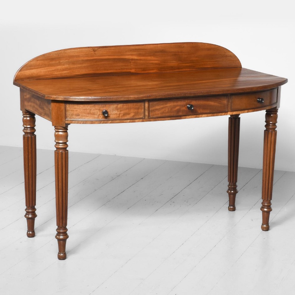 An antique Regency mahogany side table featuring a curved gallery back, three frieze drawers with small round dark knobs, and four reeded turned legs.