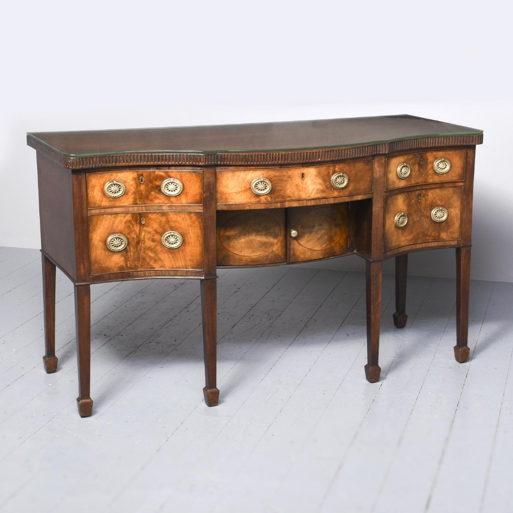 A George III serpentine front mahogany sideboard featuring brass ring-pull handles, inlaid timber detailing, and square tapered legs with spade feet.