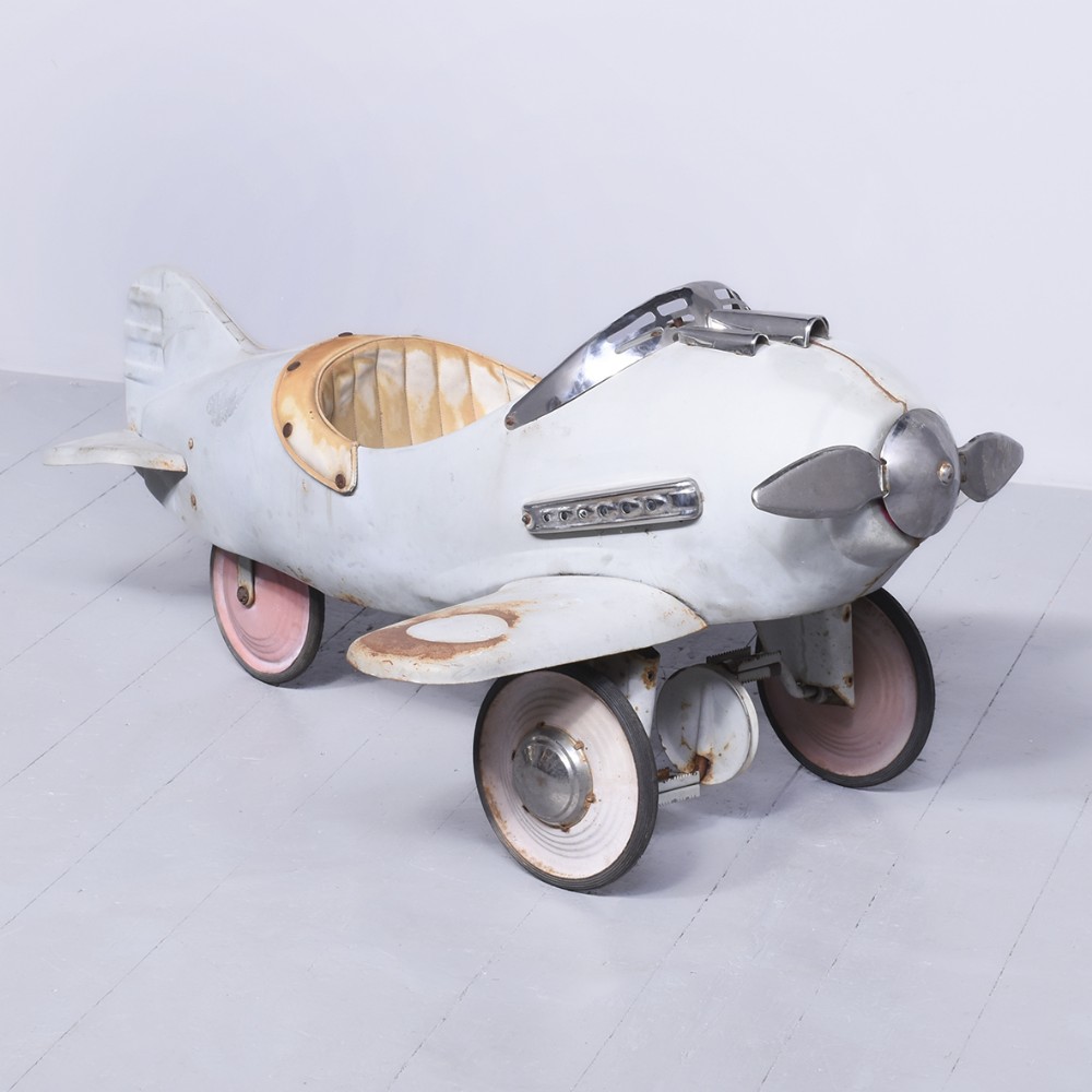 A vintage off-white metal pedal plane featuring a front propeller, chrome windshield trim, and pink-rimmed wheels.