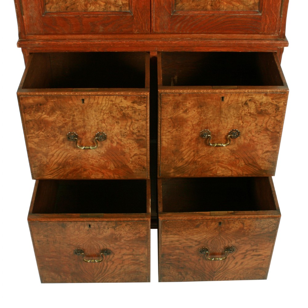 A tall two-section wooden cabinet featuring upper panelled doors with figured veneer and four lower drawers with brass handles. View VI