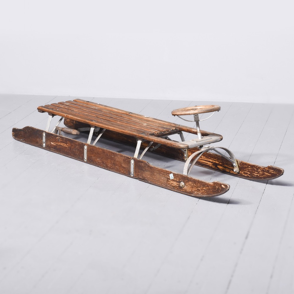 A long vintage wooden steering sledge with a slatted timber seat, a front wooden steering wheel, and curved runners reinforced with metal brackets.