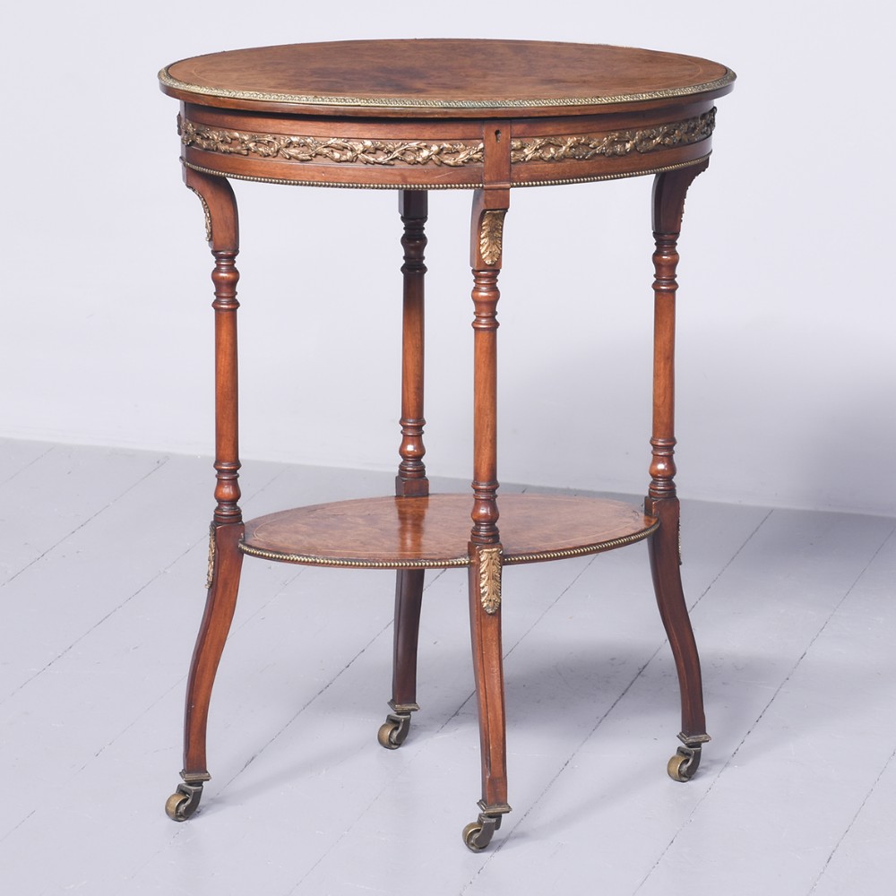 An oval Victorian amboyna occasional table with a gilt bronze frieze mount, a lower tier shelf, turned legs, and brass castors.
