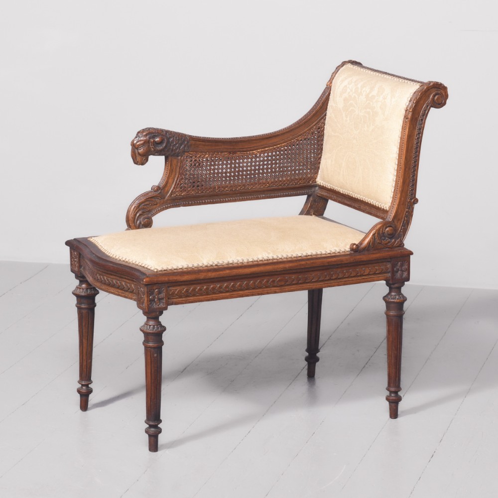 A carved walnut window seat featuring a single raised armrest topped with a carved animal head motif, a woven cane side panel, cream fabric upholstery, and fluted turned legs.