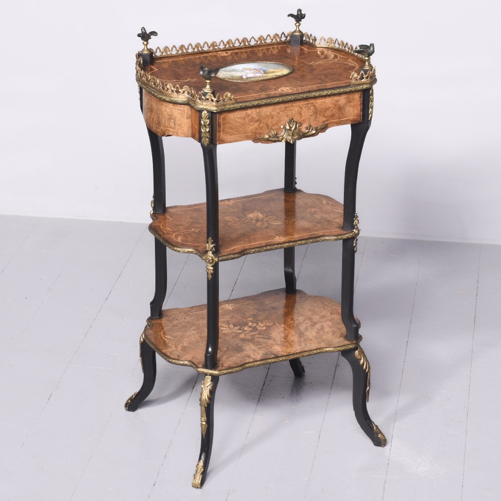 A three-tier Louis XV style etagere with burr walnut surfaces, floral marquetry, ebonised cabriole legs, brass galleries, and an inlaid oval porcelain plaque.