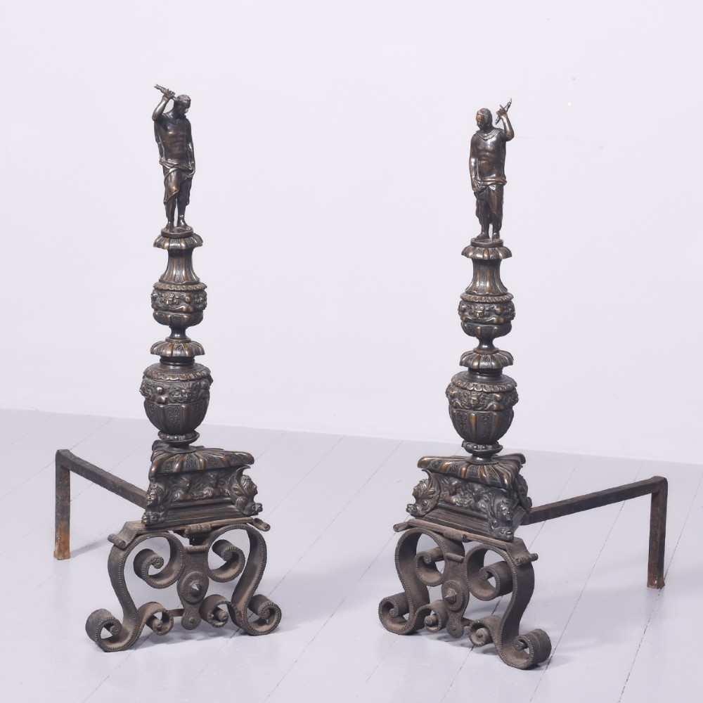 A pair of tall antique bronze and steel fire dogs featuring classical figural finials, ornate scrolled bases, and horizontal iron billet bars.