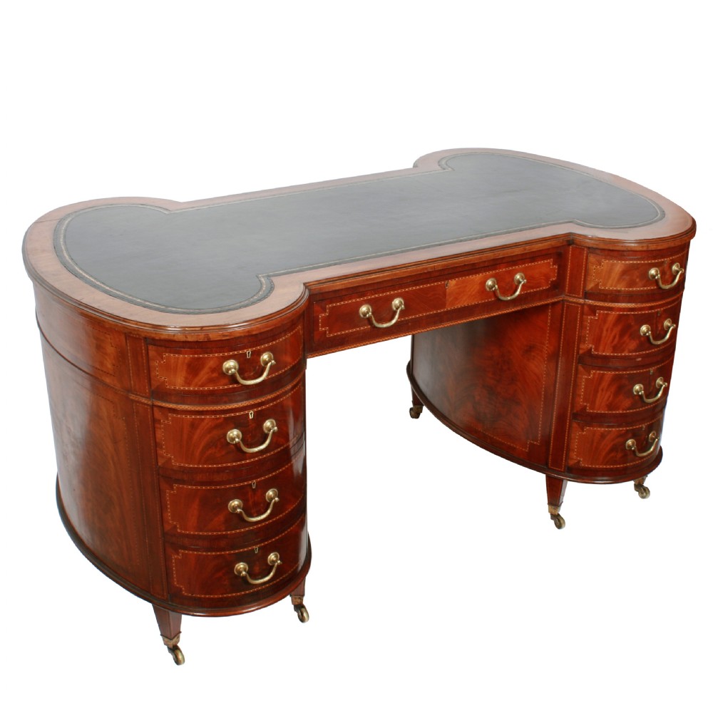An antique mahogany hourglass-shaped partners desk featuring an inset green leather top, curved drawers with brass handles, and brass castors.