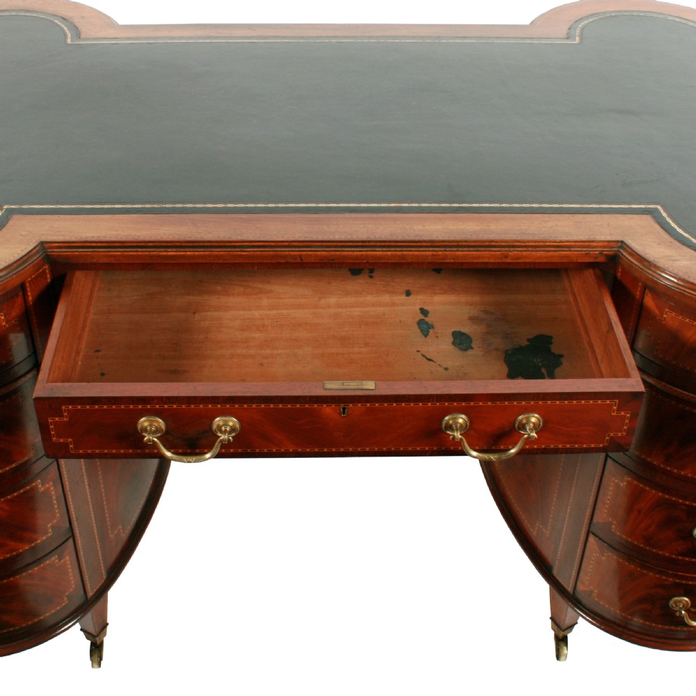 An antique mahogany hourglass-shaped partners desk featuring an inset green leather top, curved drawers with brass handles, and brass castors. View III