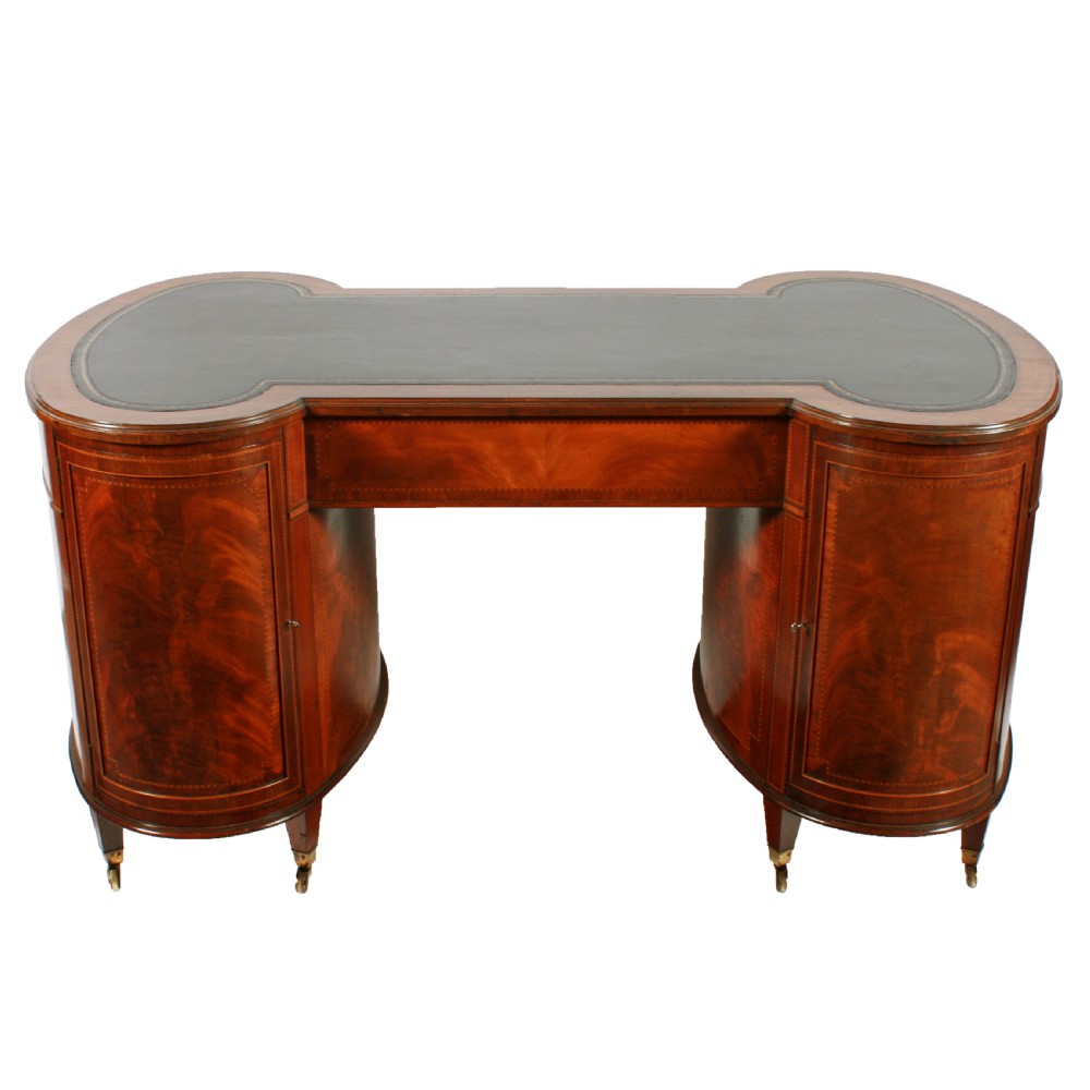 An antique mahogany hourglass-shaped partners desk featuring an inset green leather top, curved drawers with brass handles, and brass castors. View VI