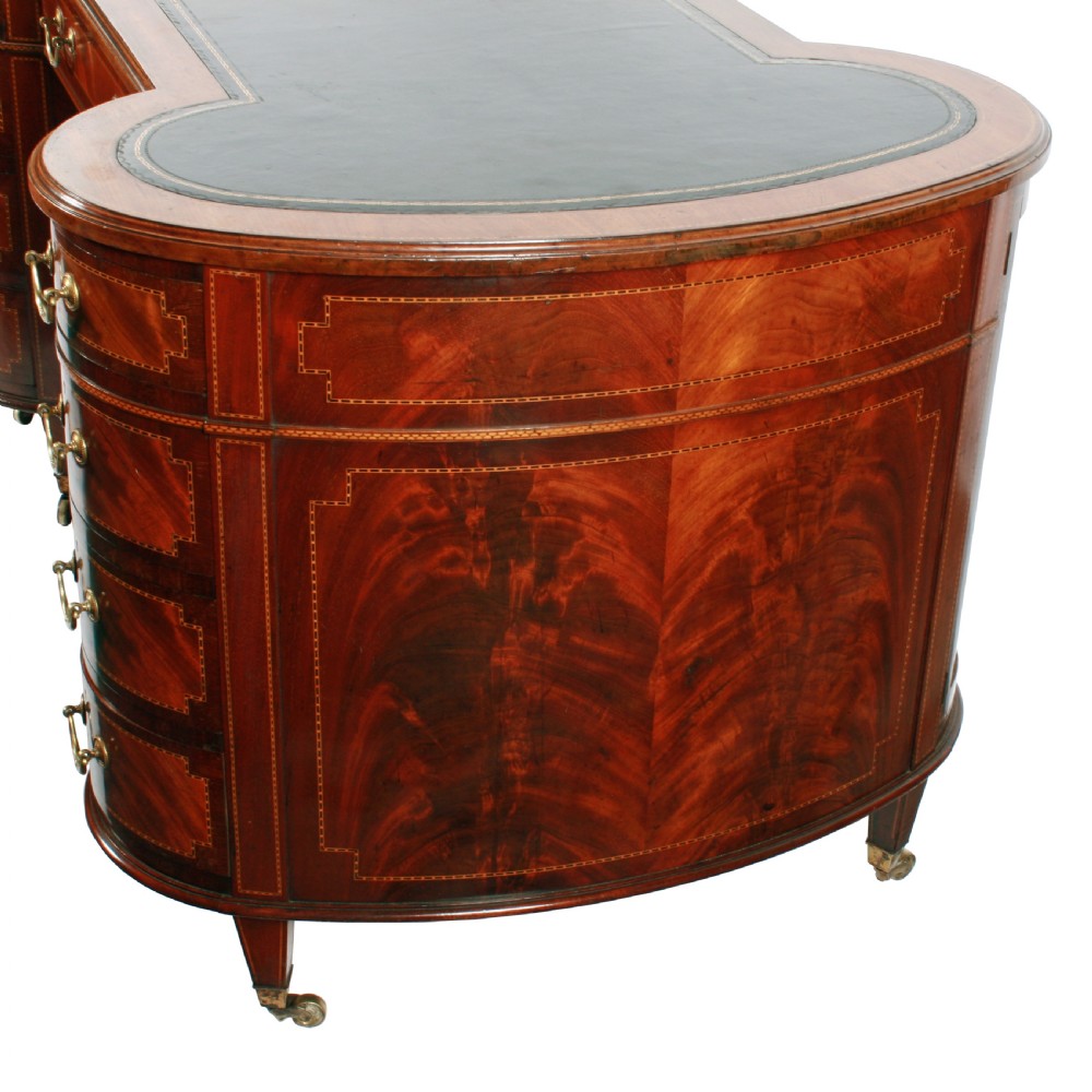 An antique mahogany hourglass-shaped partners desk featuring an inset green leather top, curved drawers with brass handles, and brass castors. View IX