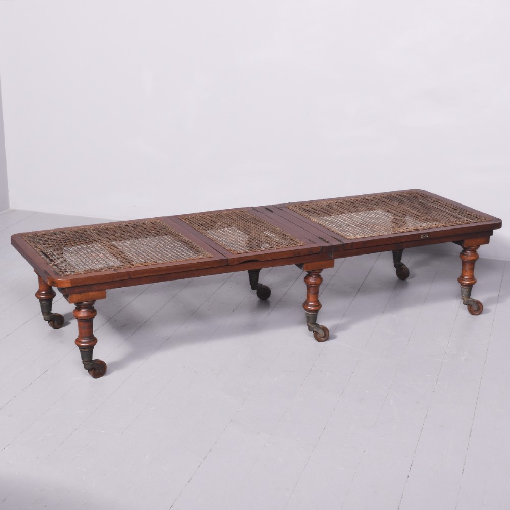 A low-profile George IV mahogany folding campaign bed featuring three caned rattan panels, turned legs, and brass castors.
