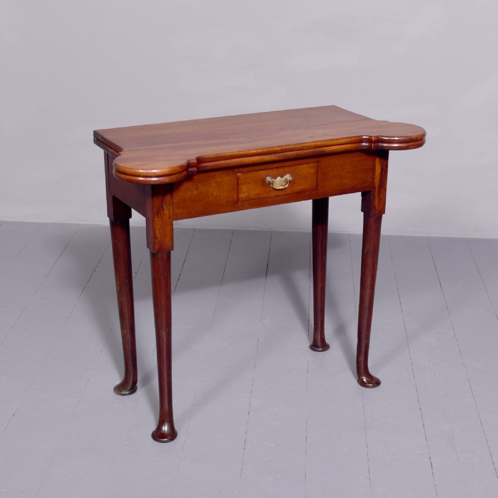 A George II mahogany fold-over games table featuring projecting rounded corners, a central drawer with brass drop handle, and slender legs ending in pad feet.