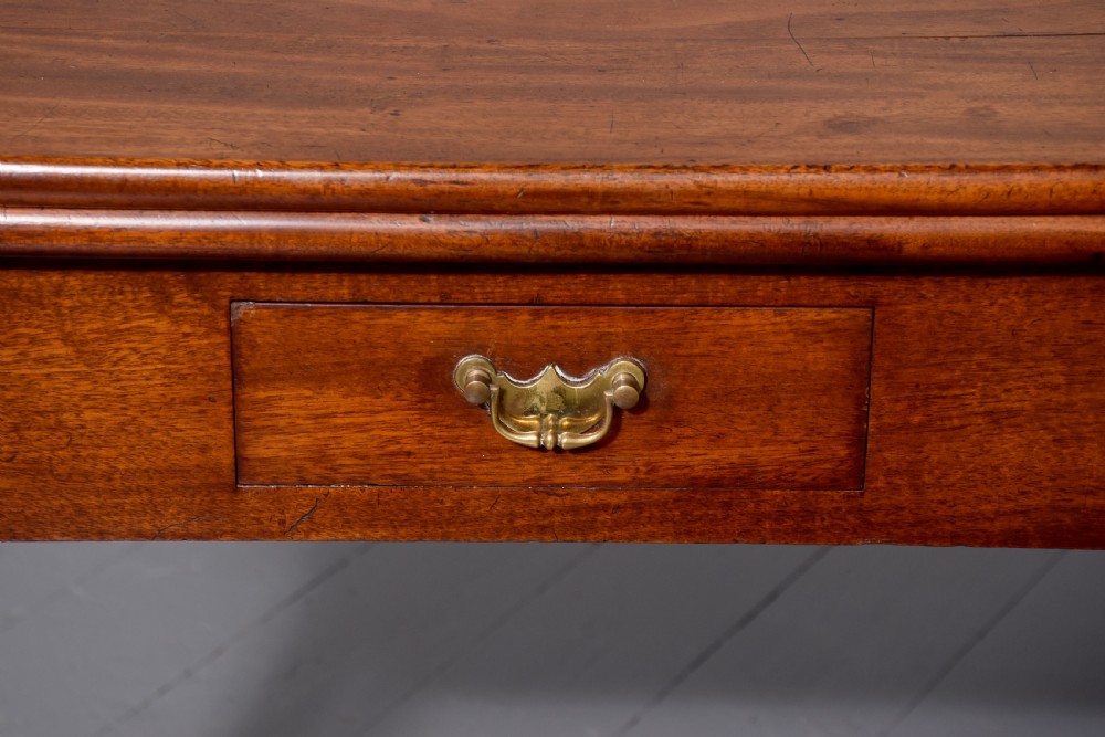 A George II mahogany fold-over games table featuring projecting rounded corners, a central drawer with brass drop handle, and slender legs ending in pad feet. View II