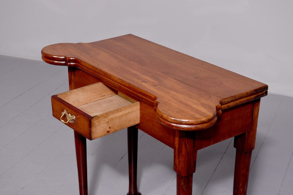 A George II mahogany fold-over games table featuring projecting rounded corners, a central drawer with brass drop handle, and slender legs ending in pad feet. View V