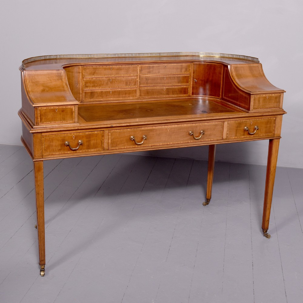 A satinwood Carlton House writing desk featuring a brass gallery railing, U-shaped superstructure with small drawers, leather writing surface, and square tapered legs on castors.