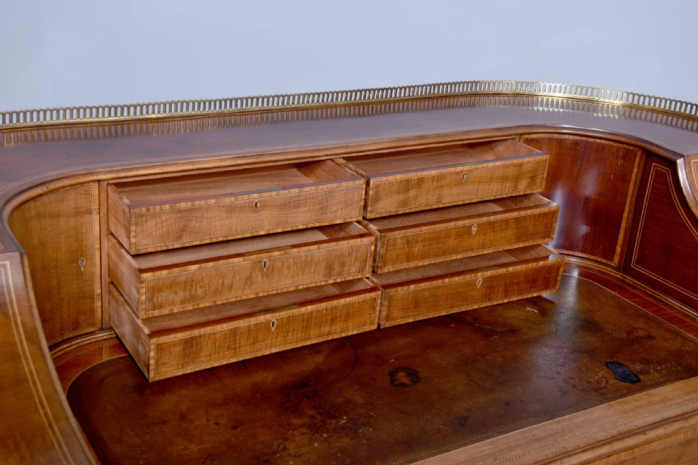 A satinwood Carlton House writing desk featuring a brass gallery railing, U-shaped superstructure with small drawers, leather writing surface, and square tapered legs on castors. View VI