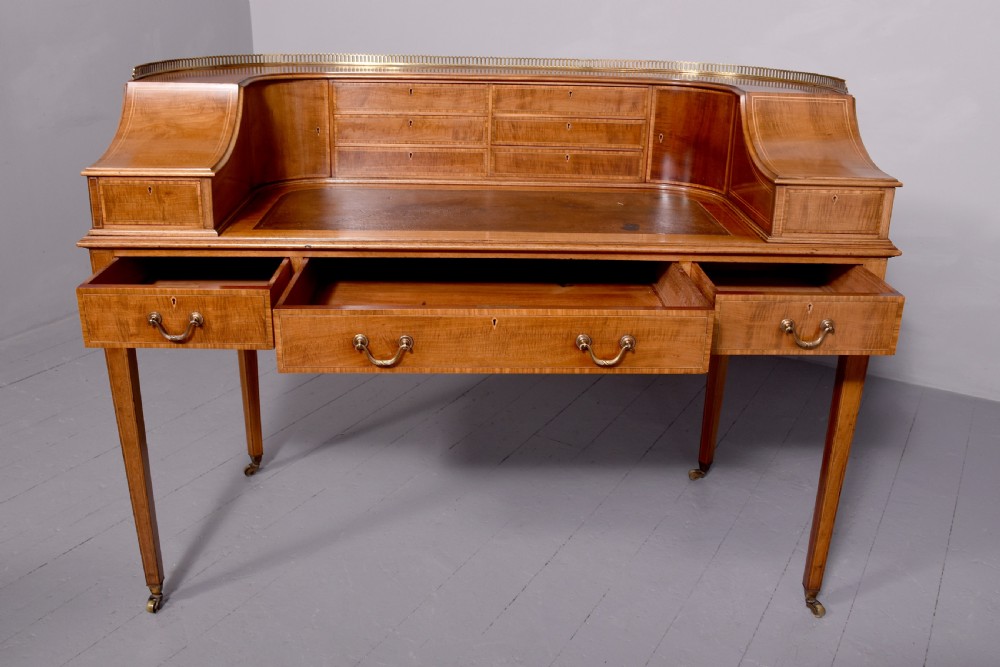 A satinwood Carlton House writing desk featuring a brass gallery railing, U-shaped superstructure with small drawers, leather writing surface, and square tapered legs on castors. View IX