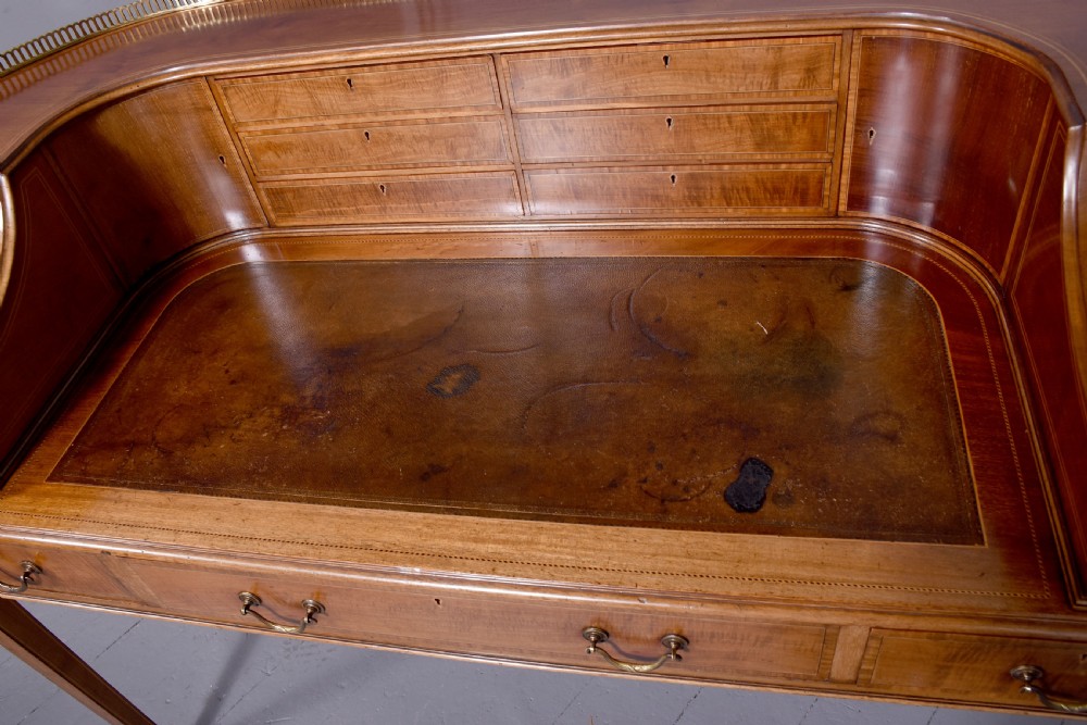 A satinwood Carlton House writing desk featuring a brass gallery railing, U-shaped superstructure with small drawers, leather writing surface, and square tapered legs on castors. View X