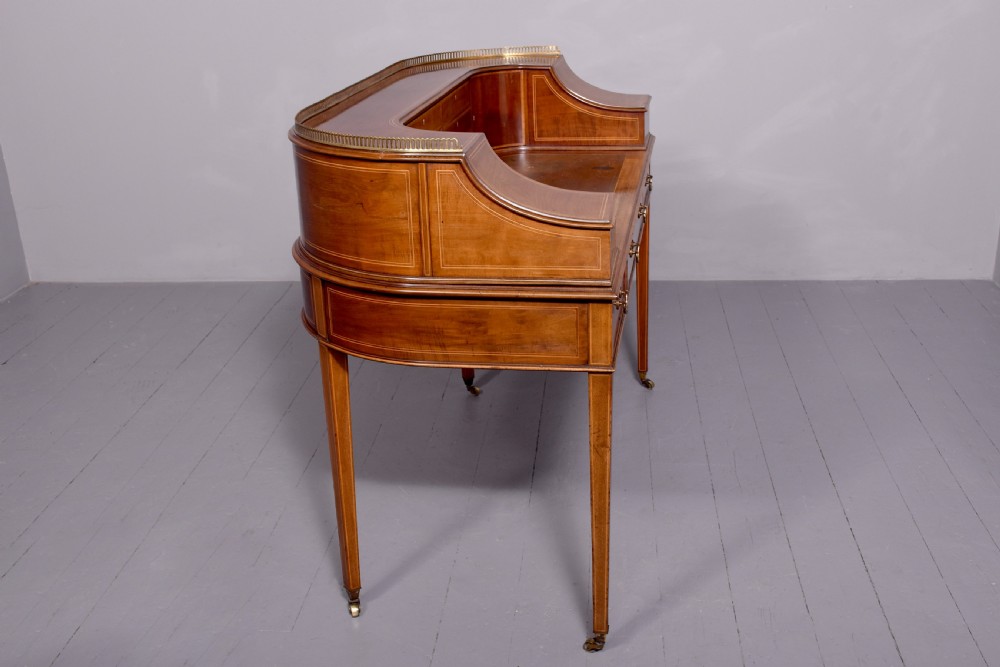 A satinwood Carlton House writing desk featuring a brass gallery railing, U-shaped superstructure with small drawers, leather writing surface, and square tapered legs on castors. View XII