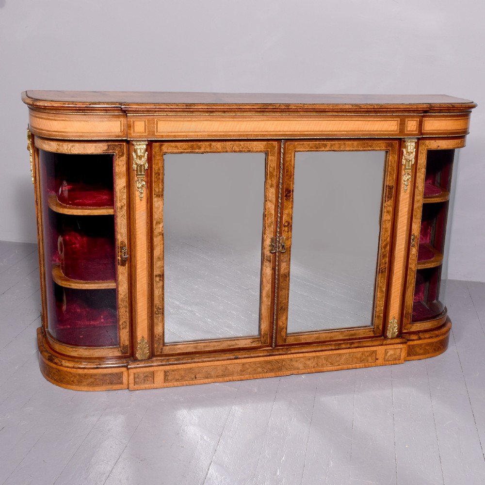 A Mid-Victorian burr walnut and satinwood credenza with mirrored central doors, curved glass side cupboards, and gilt ormolu mounts.