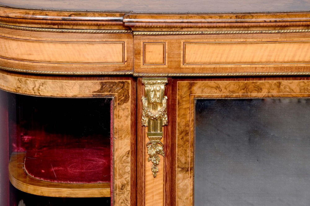 A Mid-Victorian burr walnut and satinwood credenza with mirrored central doors, curved glass side cupboards, and gilt ormolu mounts. View II