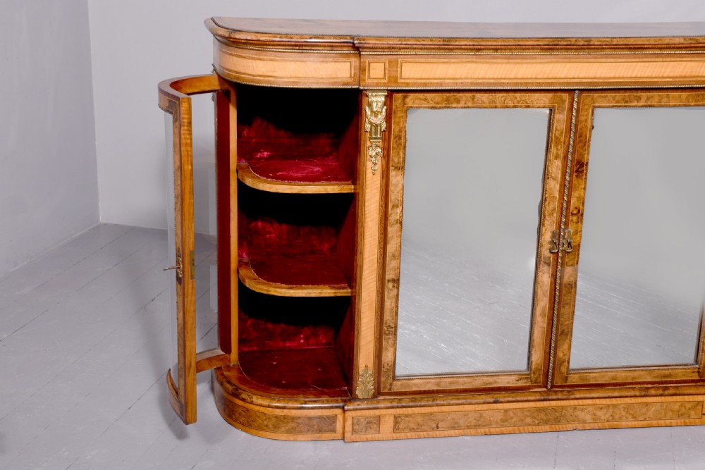 A Mid-Victorian burr walnut and satinwood credenza with mirrored central doors, curved glass side cupboards, and gilt ormolu mounts. View XIV