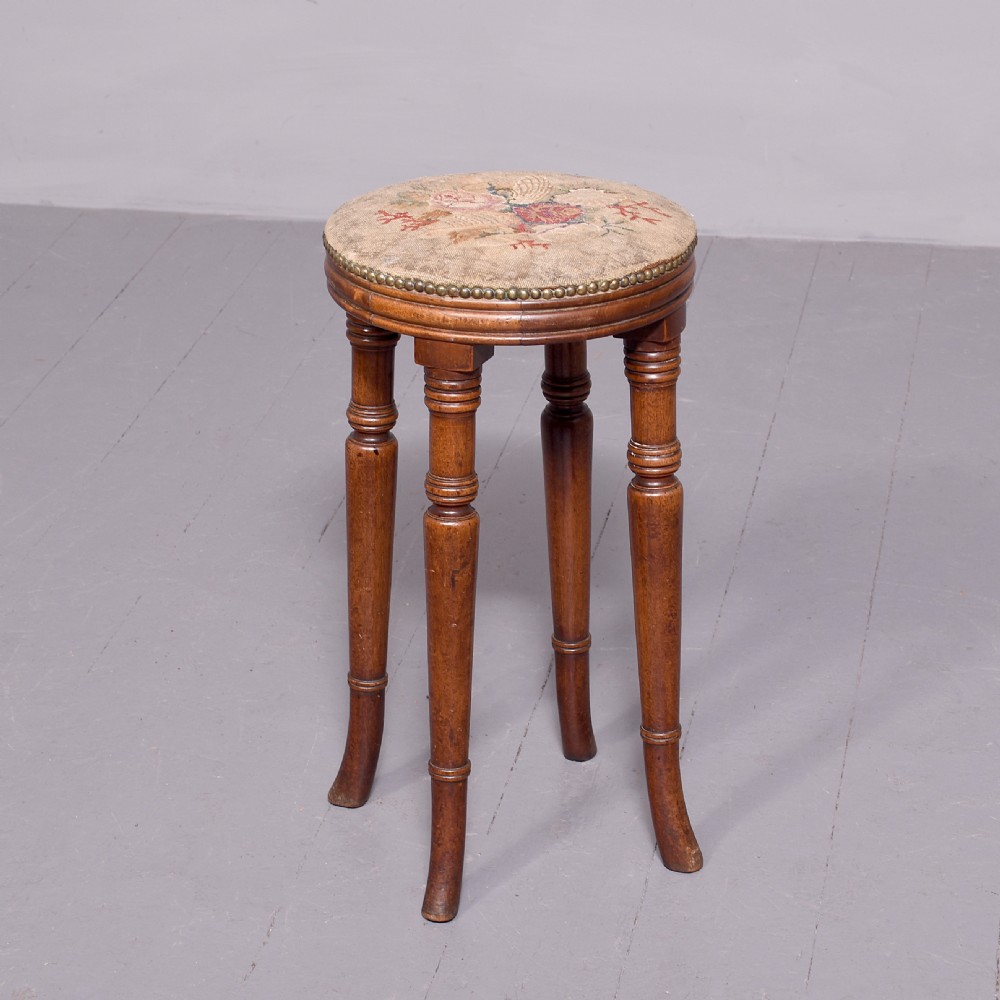 A tall George III circular wooden stool featuring four turned legs, a floral tapestry seat, and brass stud detailing along the rim.