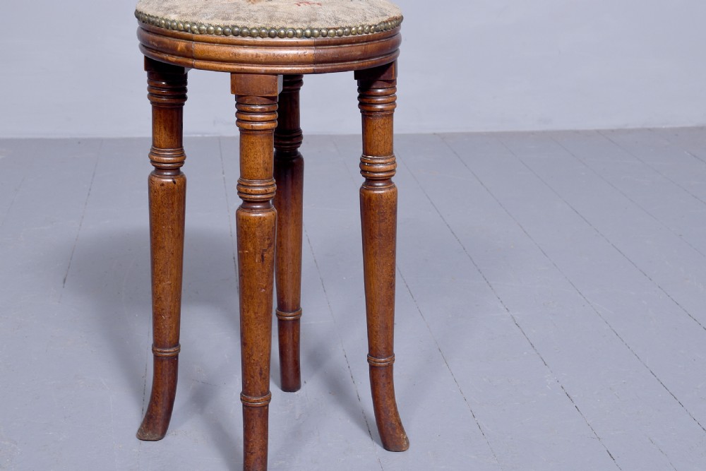 A tall George III circular wooden stool featuring four turned legs, a floral tapestry seat, and brass stud detailing along the rim. View III