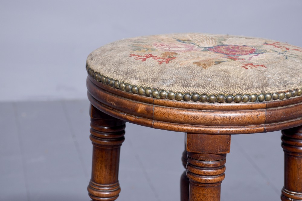 A tall George III circular wooden stool featuring four turned legs, a floral tapestry seat, and brass stud detailing along the rim. View IV
