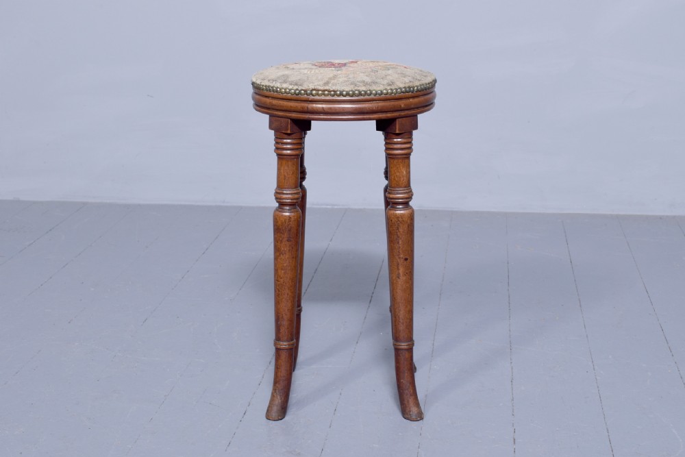 A tall George III circular wooden stool featuring four turned legs, a floral tapestry seat, and brass stud detailing along the rim. View V
