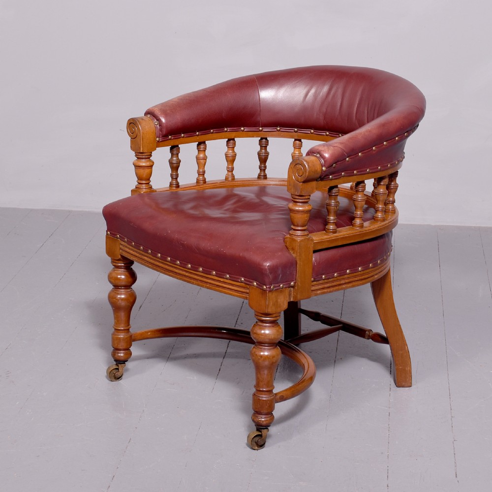 A Victorian walnut desk chair featuring a curved burgundy leather backrest with brass stud detailing, turned spindles, and front turned legs on brass castors.