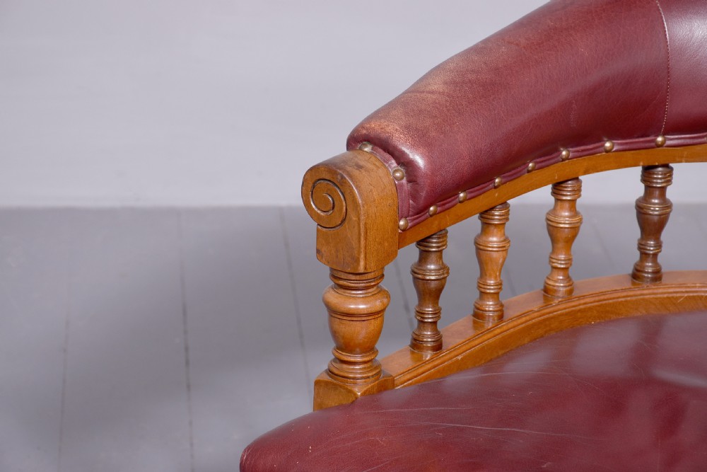 A Victorian walnut desk chair featuring a curved burgundy leather backrest with brass stud detailing, turned spindles, and front turned legs on brass castors. View II