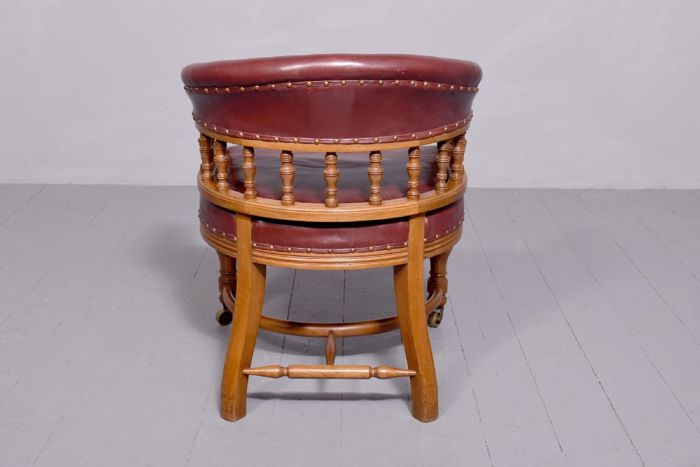 A Victorian walnut desk chair featuring a curved burgundy leather backrest with brass stud detailing, turned spindles, and front turned legs on brass castors. View V