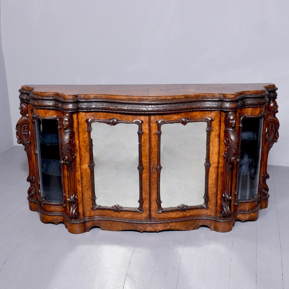 A mid-Victorian burr walnut serpentine-fronted credenza featuring carved female figure pilasters, central mirrored doors, and curved glass side display cabinets.