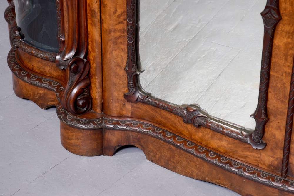 A mid-Victorian burr walnut serpentine-fronted credenza featuring carved female figure pilasters, central mirrored doors, and curved glass side display cabinets. View V