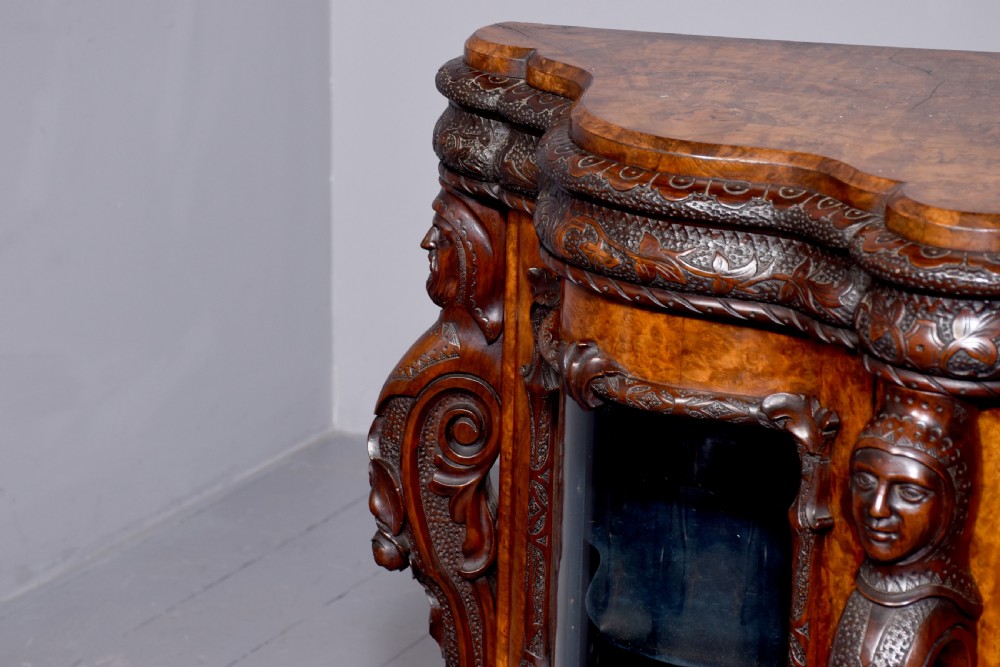 A mid-Victorian burr walnut serpentine-fronted credenza featuring carved female figure pilasters, central mirrored doors, and curved glass side display cabinets. View VI