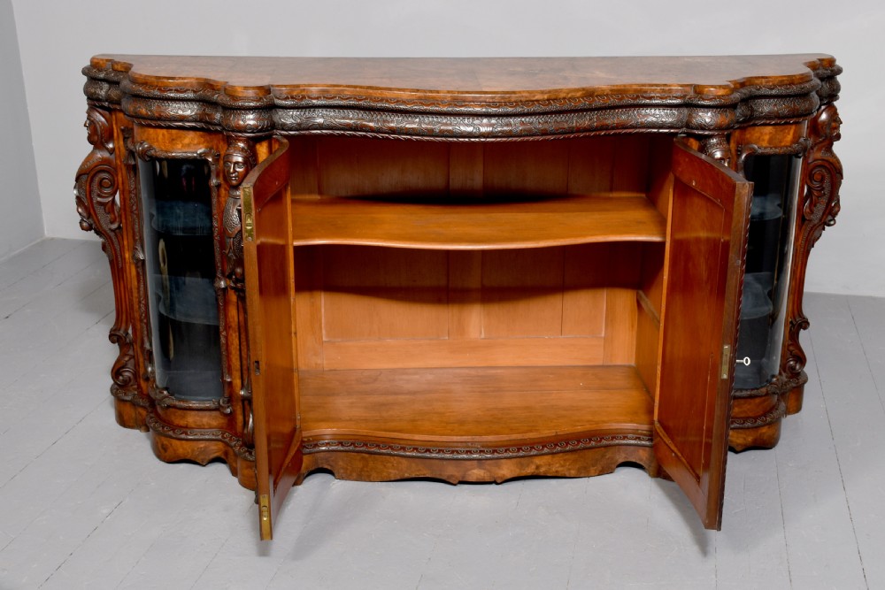 A mid-Victorian burr walnut serpentine-fronted credenza featuring carved female figure pilasters, central mirrored doors, and curved glass side display cabinets. View X