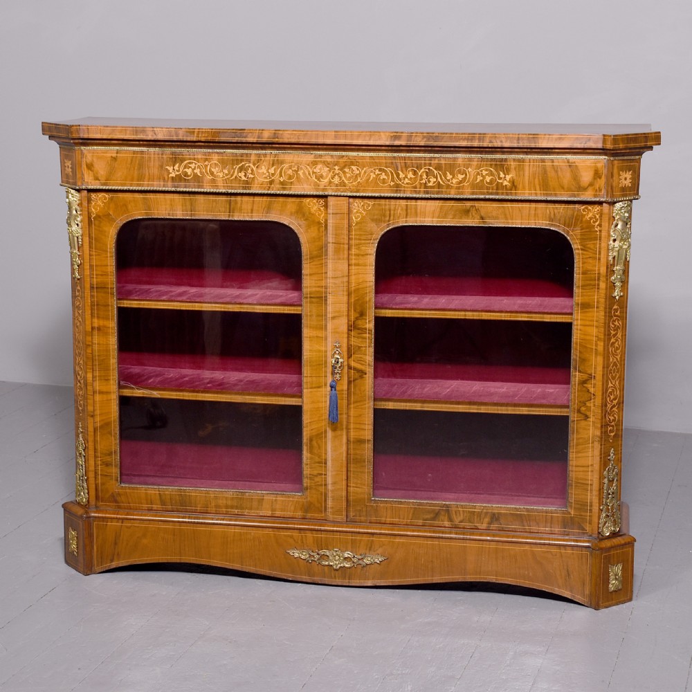 A Victorian figured walnut cabinet featuring twin glazed doors, intricate marquetry floral inlay along the frieze, gilt bronze mounts, and a red fabric-lined interior with three shelves.