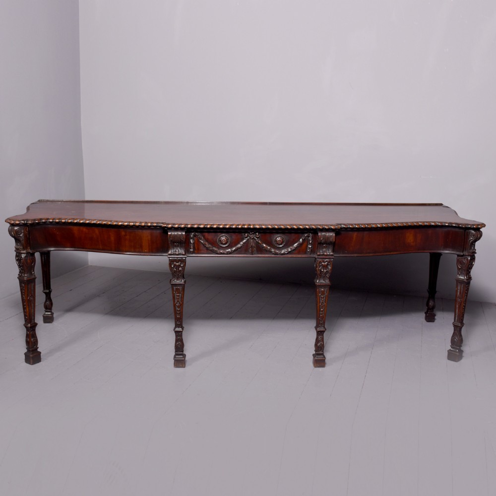 A wide George III dark mahogany serving table supported by six carved legs with intricate drapery swags running across the front frieze.