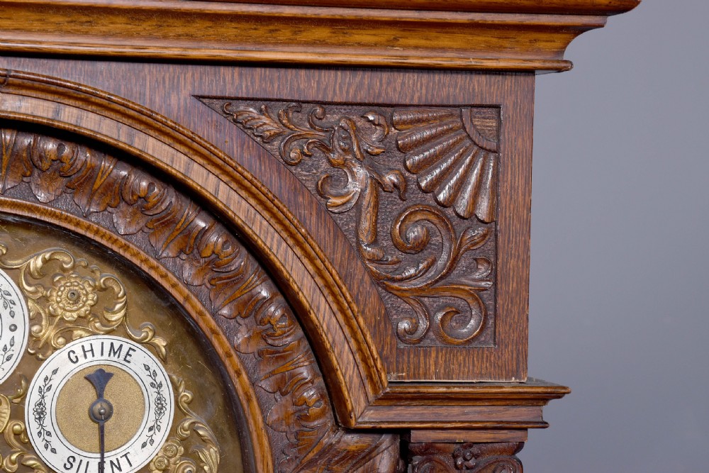 A late Victorian tall carved oak grandfather clock with a swan-neck pediment, arched brass dial, and extensive foliate relief carvings on the body and base. View V