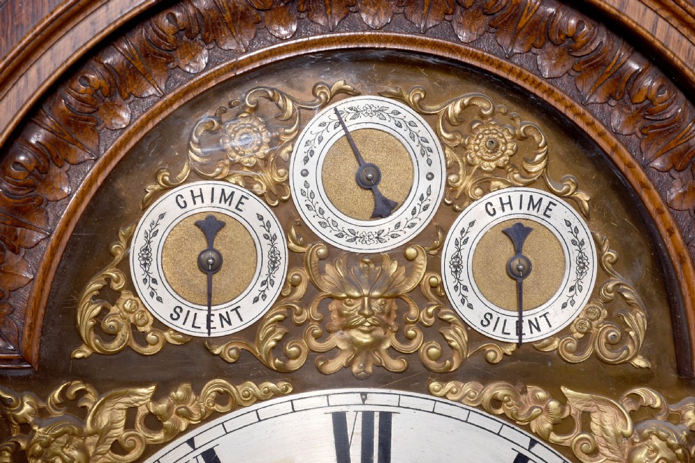 A late Victorian tall carved oak grandfather clock with a swan-neck pediment, arched brass dial, and extensive foliate relief carvings on the body and base. View VI
